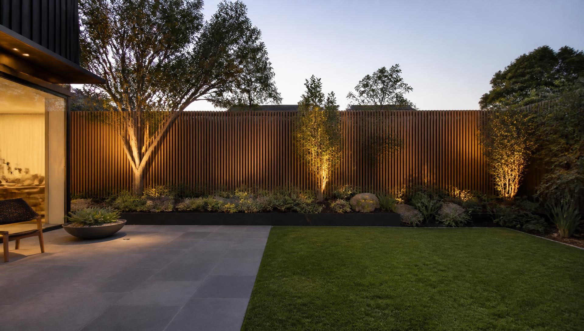 Contemporary Melbourne backyard with bluestone paving, lawn and lit garden beds at dusk